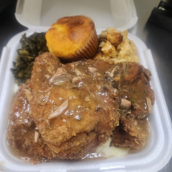 Fried chicken with gravy, collard greens, cornbread, and macaroni and cheese in a takeout container.