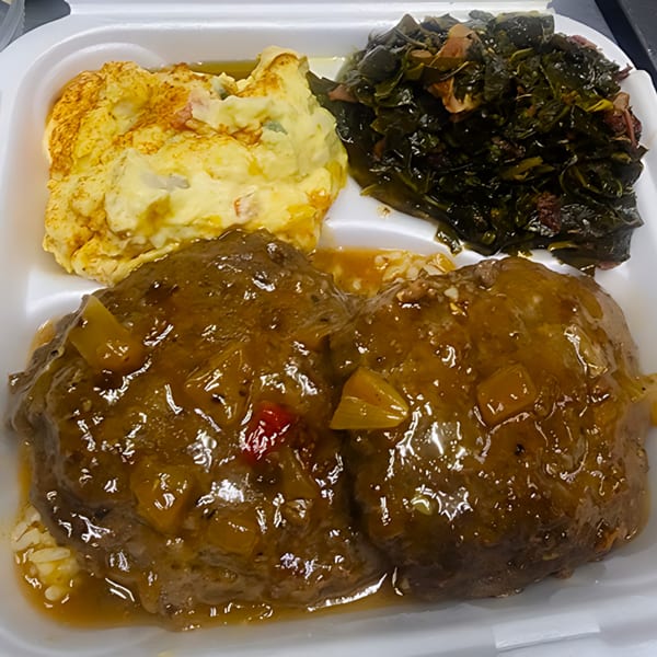 Soul food meal with two Salisbury steaks, mashed potatoes, and collard greens in a takeout container.